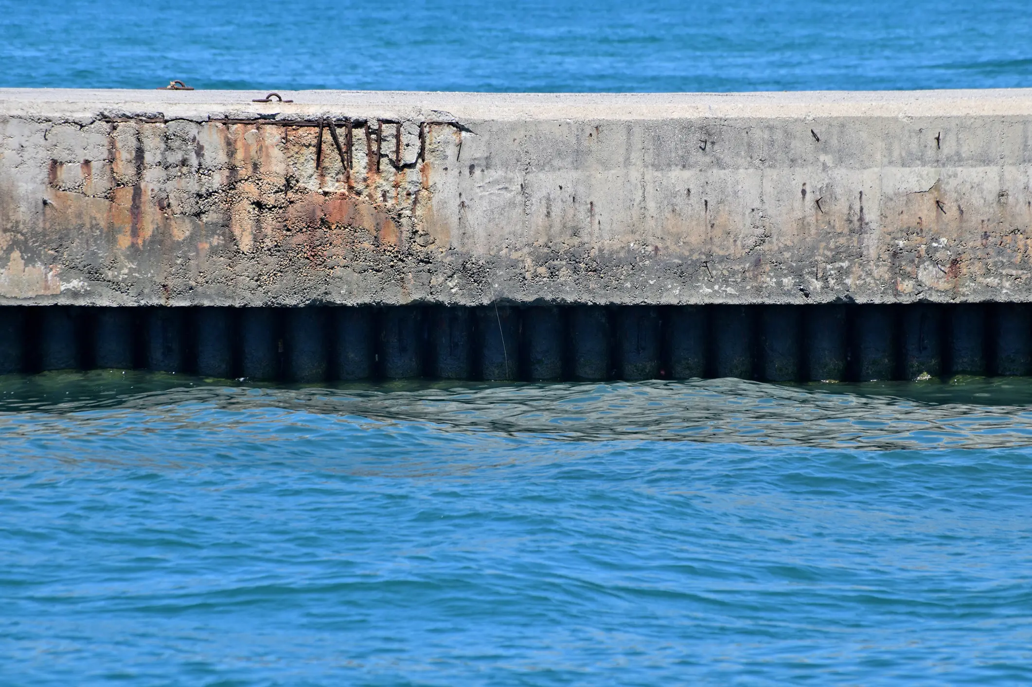 A fragment of a reinforced concrete berth at the base of which iron piles are visible. Blue sea in front of and behind the breakwater.