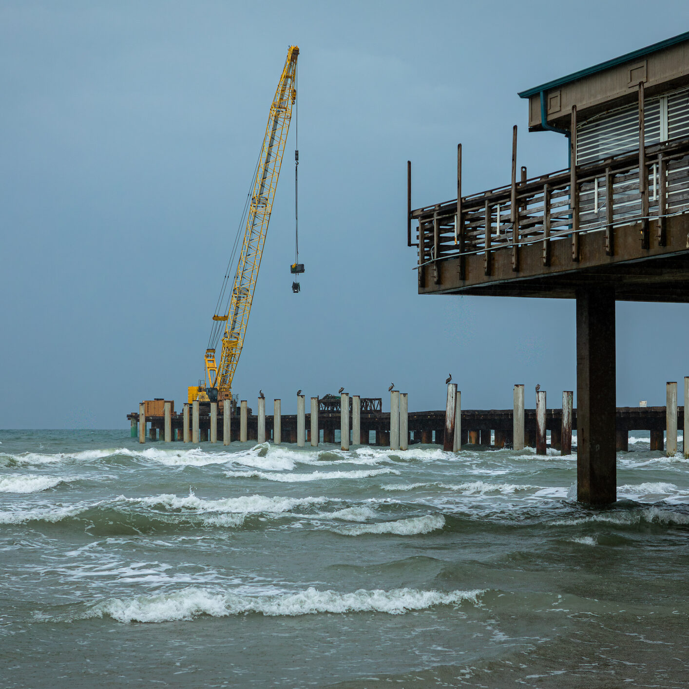 Yellow crane restoring a pier over rough ocean waves on a stormy day at the Texas coast. Industrial meets natural seascape.